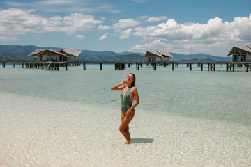 Young slim tourist woman enjoy suntan, standing in the tourquise water in front of wooden owrwater bungalows, wearing swimsuit. Vacation concept