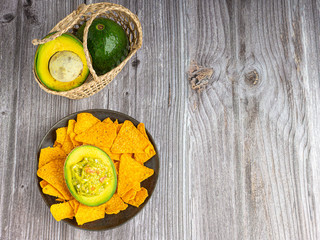 Guacamole with corn tortilla chips in plate and avocados in basket on wooden table