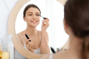 Smiling young woman looking at mirror, using mascara