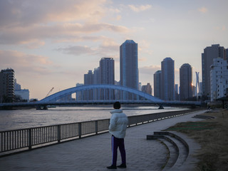 Fototapeta na wymiar young man in puffer jacket standing in front of skyline and river in tokyo at sunset