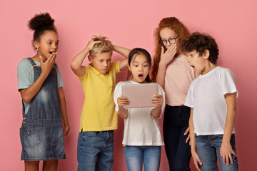 Obraz premium Surprised boys and girls looking at tablet computer together, learning shocking news over pink background