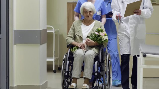 Tilt up shot of elderly female patient holding bouquet of flowers and riding on wheelchair through hospital corridor with assistance of female nurse while male doctor on lab coat walking beside them