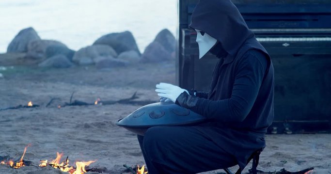 a man in a venetian mask on the seashore plays the glucophone against the background of a burning old piano