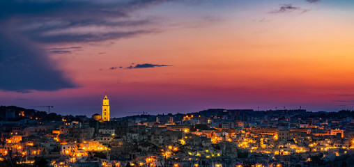 The spectacular fiery sunset and the panorama of the Sassi di Matera in Basilicata, Italy. The top view of the city from the park of the Rupestrian Churches. The lights come on as night falls.
