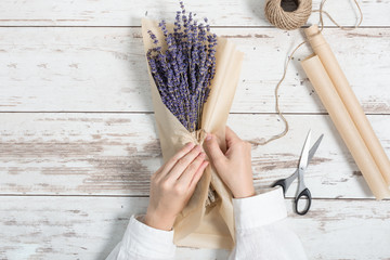 Woman wrapping dried lavender flowers bunch in craft wrapping paper on white wooden background top view.