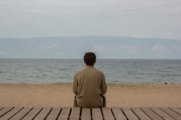 Young guy resting and meditating on a wooden deck in front of a sandy beach looking into the sea