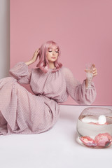 Girl with dyed pink hair in long dress sitting on the floor with a round aquarium. Portrait woman with hair coloring at pink wall. Perfect hairstyle and hair styling