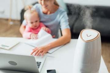 Selective focus of humidifier on table near woman holding baby girl and working at home - obrazy, fototapety, plakaty