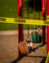 Empty saddle style swings at a playground covered in yellow caution tape written in english and spanish.