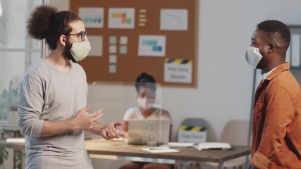 Afro-American and Arab male colleagues in face masks greeting each other with elbow bump and chatting while working in office during coronavirus outbreak - Powered by Adobe