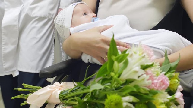 Close up view of hands of mother holding cute newborn baby with soother and flower bouquet while riding wheelchair through maternity hospital