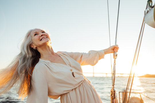 Beautiful mature woman with long hair enjoying sunset. Smiling woman holding a rope and looking up while standing on a sailboat.