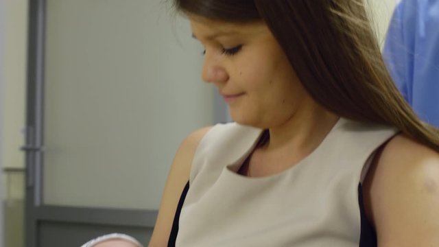 Close up shot of happy mother holding newborn baby with soother on arms and looking at him with love while riding wheelchair with assistance of nurse in maternity hospital 