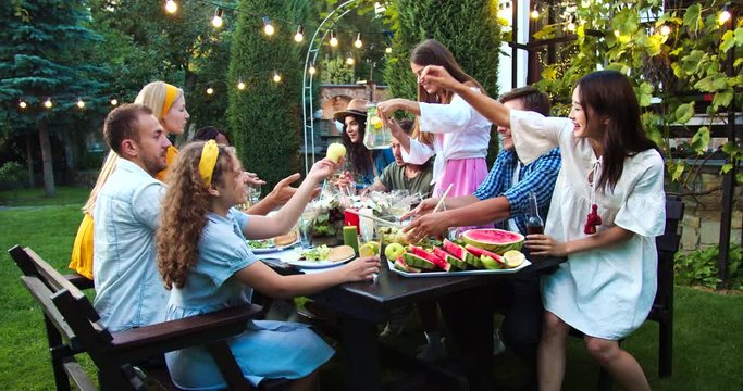 Young people eating and drinking at garden party. African American and Caucasian girls speaking. Happy guys passing dishes. Asian female clinks glasses with friend. Celebration concept