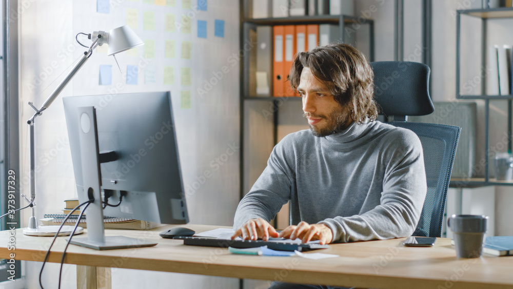 Poster handsome long haired entrepreneur sitting at his desk in the office works on desktop computer, worki