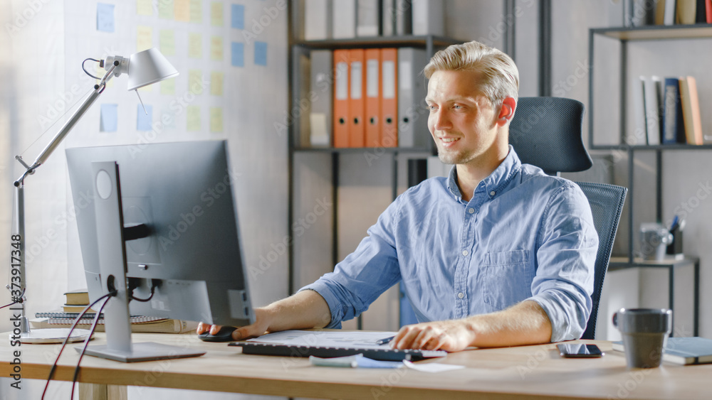 Poster handsome blonde businessman sitting at his desk in the office works on desktop computer, working wit