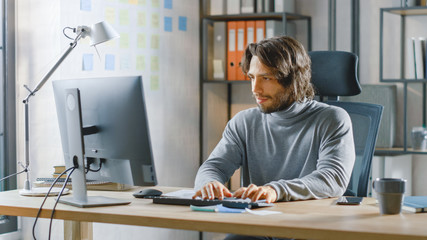 Handsome Long Haired Entrepreneur Sitting at His Desk in the Office Works on Desktop Computer, Working with Documents, Charts, Graphs, Statistic and Strategy. Creative Entrepreneur Using Computer