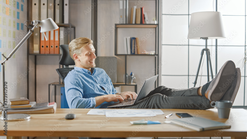 Poster handsome young entrepreneur works on a laptop with his legs up on the desk. designer uses notebook t