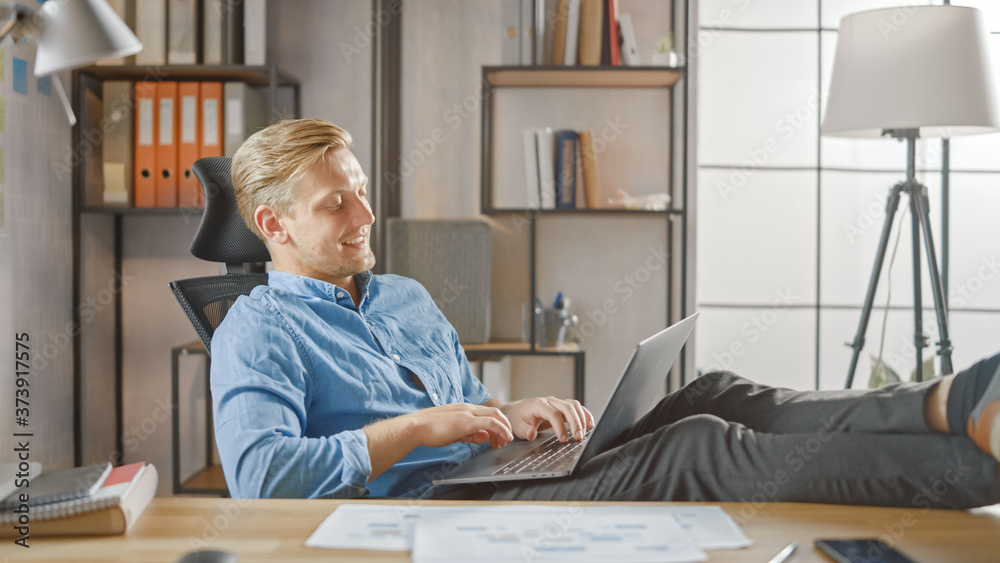 Poster handsome young entrepreneur works on a laptop with his legs up on the desk. designer uses notebook t