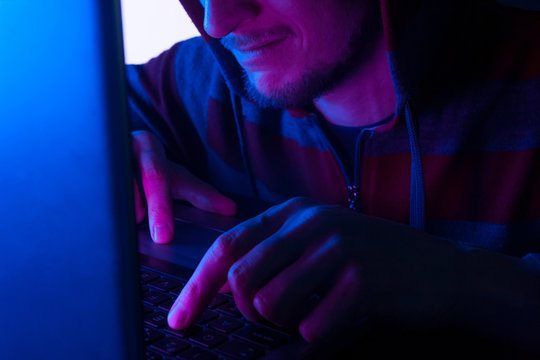 smiling young hacker in hoodie typing on keyboard, hacking personal data concept