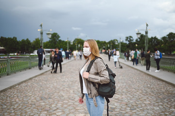 Young blonde woman walking in city street wearing face protective mask for Covid 19 prevention. Caucasian young student or traveler tourist among the crowd with backpack. Corona virus concept