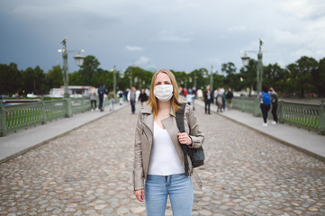 Young blonde woman walking in city street wearing face protective mask for Covid 19 prevention. Caucasian young student or traveler tourist among the crowd with backpack. Corona virus concept