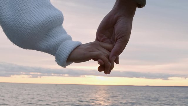 Close up of unrecognizable man and woman taking each other hand while standing on seashore at sunset
