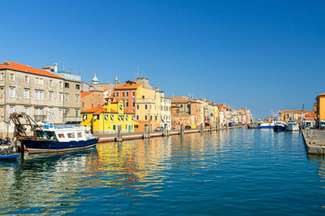 Fototapeta na wymiar Narrow water canal with boats and fishing ships moored near embankment and colorful buildings in Chioggia town historical centre, blue sky background in summer day, Veneto Region, Northern Italy