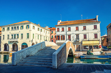Obraz premium Stone brick bridge Ponte Pescheria across Vena water canal and old buildings in historical centre of Chioggia town, blue sky background in summer day, Veneto Region, Northern Italy