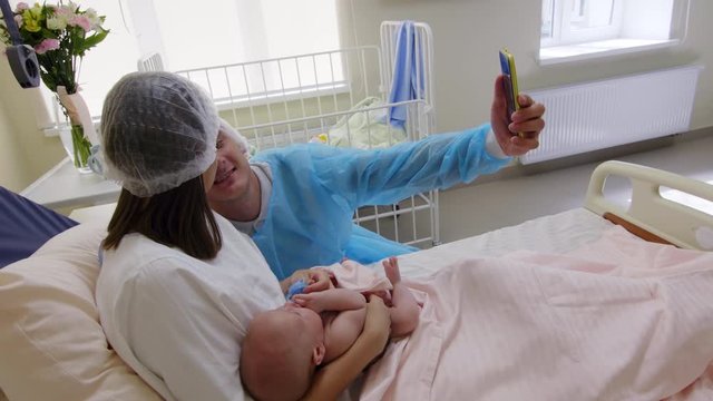 Happy family couple kissing and using smartphone while taking selfie with newborn baby in maternity hospital ward