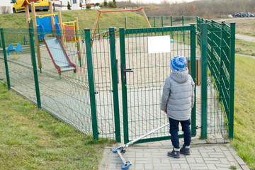 View of a child with a scooter standing in front of a closed playground, child reading the inscription on the plate, quarantine