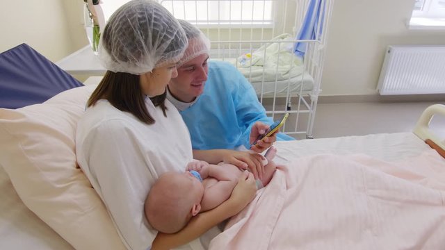 Cheerful wife and husband in medical hats holding newborn baby and talking a selfie with smartphone in maternity hospital ward