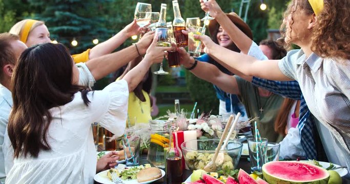 Multi-ethnic happy young people gathered together outdoor. Male and female joyful friends eating and drinking at table in garden. Youth in good mood raising glasses and clinking. Summer party concept