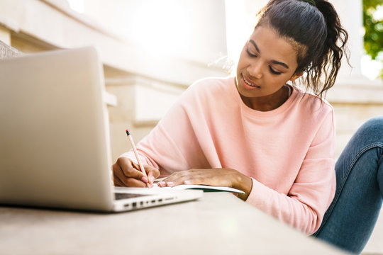 Image of pleased african american student girl doing homework with laptop