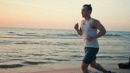 Sporty Man is Jogging on Beach with Phone and Running along Sea Coast at Sunset