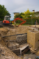 Fototapeta na wymiar A sewer well trench and a compact crawler excavator with a rotating house platform and continuous caterpillar track on a sewer replacement work site in north west Italy 