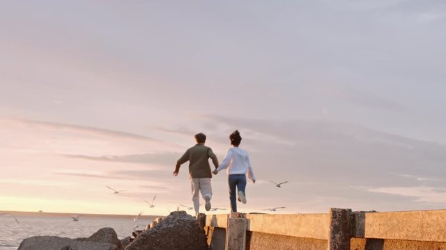 Back view of young man and woman holding hands of each other are running along pier on seashore at sunset. Gulls are flying away