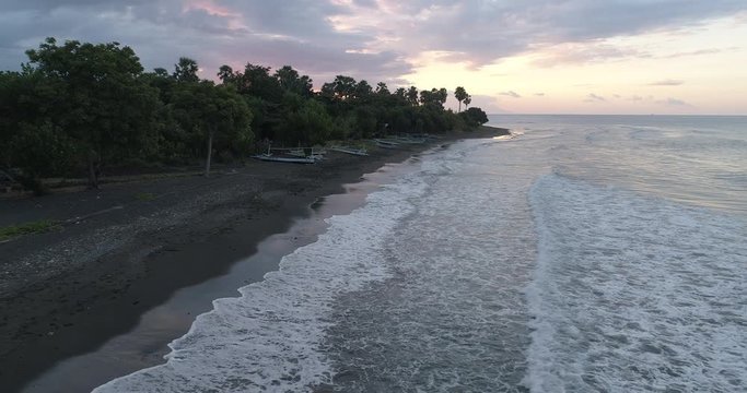 Drone shot flying backwards at low altitude over waves breaking on a black sandy beach at sunset