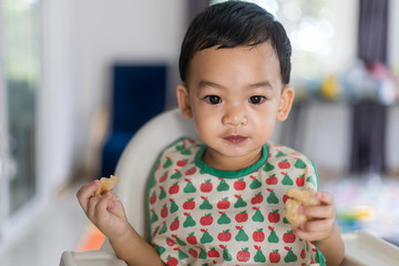 An Adorable toddler Asian baby boy (1-year-old) sitting on high feeding chair eating homemade food and learning to use hands at home.