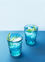 Two beautiful vintage blue glasses with cold drink and ice cubes, with fresh green mint leaves on deep blue and light blue background. Backdrop for your design.