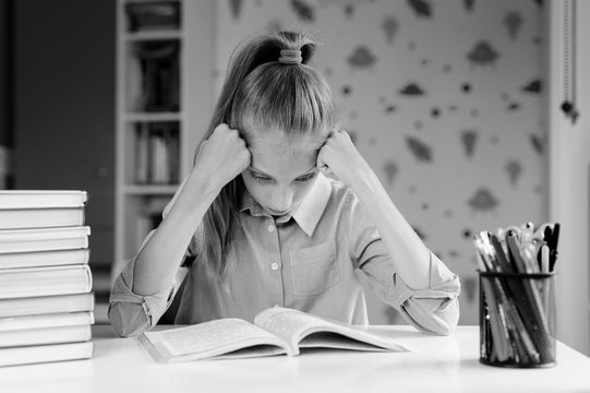 The schoolgirl in a bad mood rests her head on her hands and looks at the textbook. The girl is preparing for the exam. The girl does not want to read the book. Black and white photography.