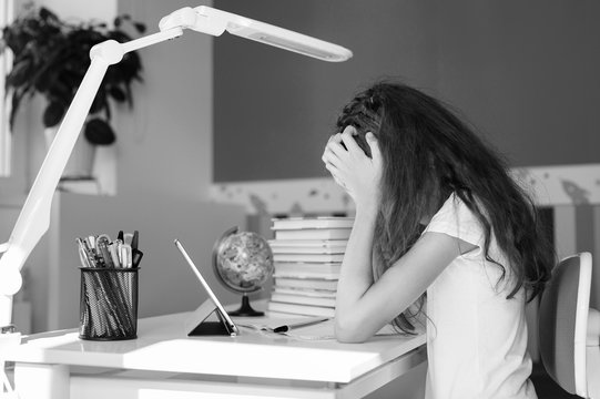  The schoolgirl in a bad mood rests her head on her hands and looks at the textbook. The girl is preparing for the exam.  Black and white photography.