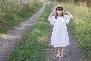 Deurstickers Mooi meisje in een hoed staat op een weg in de buurt van roggeveld. Klein meisje houdt haar hoed vast © Albert Ziganshin