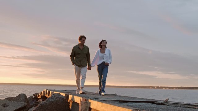 Wide shot of young man and woman holding hands of each other are walking along pier on seashore at sunset and enjoying the view