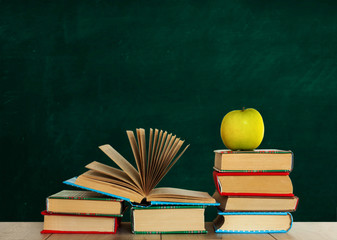 Open textbook, pile of books in colorful covers and green apple on wooden table with green blackboard background. Distance home education. Back to school, quarantine concept of stay home.