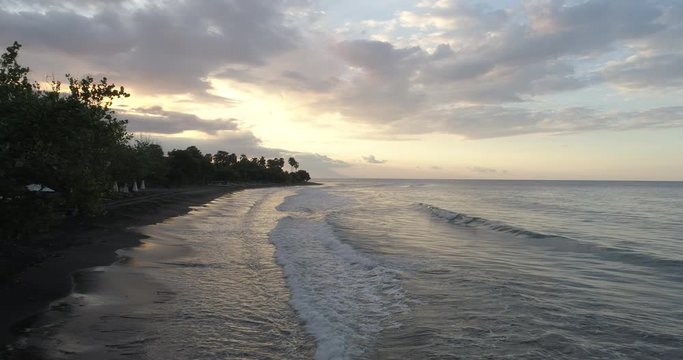 Drone shot flying over waves along a black sandy beach and ascending in a crane shot to reveal a beautiful sunset