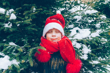 A beautiful boy in a red hat and scarf stands under a Christmas tree in the snow in winter.