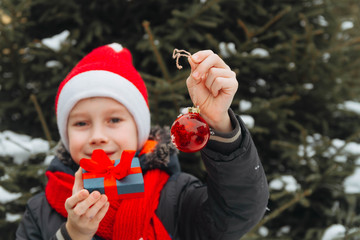 A teenage European boy in A red Santa Claus costume gives a gift for New year and Christmas. The child wants to decorate the Christmas tree with a red ball.