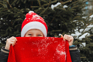 Obraz premium A teenage European boy in a red Santa suit hides behind a bright festive knitted scarf. Happy face of a child close-up. Background for Christmas.