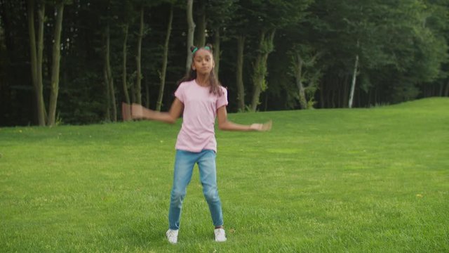 Lovely preadolescent african american girl with trendy hairstyle dancing hip-hop on green grass, making improvisation and moving to the groove happily, practicing street dance and freestyle outdoors.